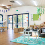 A spacious early learning room decorated with hanging branches, bunting, and paper leaf garlands, featuring a sofa, toy shelves, and large glass doors opening to the outdoors