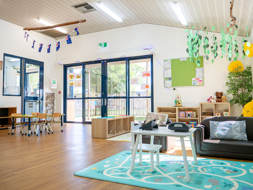 A spacious early learning room decorated with hanging branches, bunting, and paper leaf garlands, featuring a sofa, toy shelves, and large glass doors opening to the outdoors