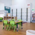 A bright early learning classroom with green chairs around a activity table, children's lockers, and a wooden dollhouse in the corner