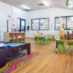 An early learning classroom with a train table on an Indigenous-design rug, green chairs at activity tables, children's artwork displayed on the walls, and dinosaur toys