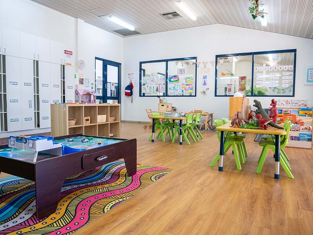 An early learning classroom with a train table on an Indigenous-design rug, green chairs at activity tables, children's artwork displayed on the walls, and dinosaur toys