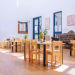 A sunny early learning dining room with timber tables and chairs, potted plants as centrepieces, an ornate sofa, and Indigenous artwork on the wall