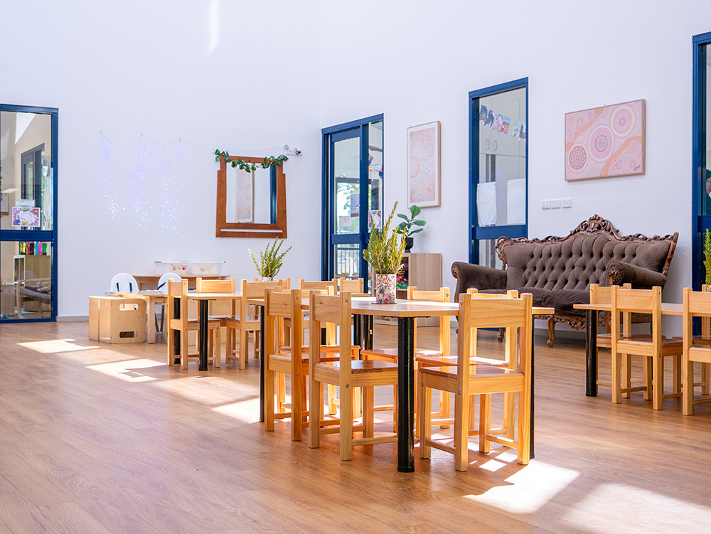 A sunny early learning dining room with timber tables and chairs, potted plants as centrepieces, an ornate sofa, and Indigenous artwork on the wall