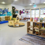 Baby room in early learning center with soft play areas, wooden climbing structure, sensory water table, ocean-themed wall decorations, organized storage cubbies with age-appropriate toys, and large windows providing natural light