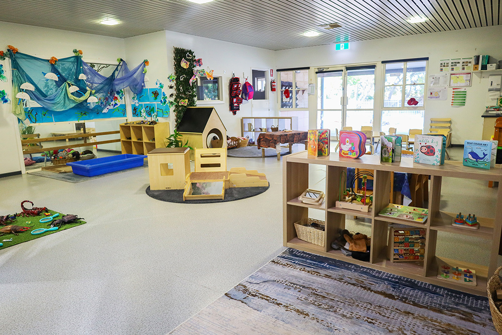 Baby room in early learning center with soft play areas, wooden climbing structure, sensory water table, ocean-themed wall decorations, organized storage cubbies with age-appropriate toys, and large windows providing natural light