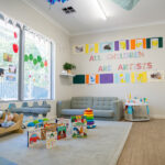 A baby and toddler room with a "All Children Are Artists" wall display, children's artwork, a soft sofa, board books on the floor, and a wooden toy cot
