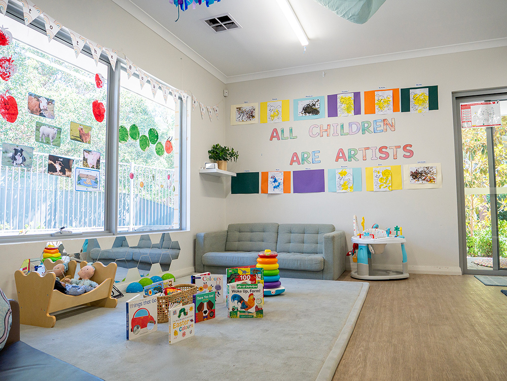A baby and toddler room with a "All Children Are Artists" wall display, children's artwork, a soft sofa, board books on the floor, and a wooden toy cot