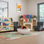 A cosy early learning reading nook with two grey armchairs, a decorative fireplace, a well-stocked children's bookshelf, and a patterned rug