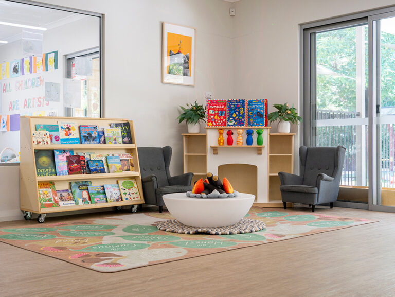 A cosy early learning reading nook with two grey armchairs, a decorative fireplace, a well-stocked children's bookshelf, and a patterned rug