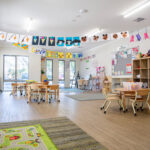 A bright early learning classroom with children's artwork strung across the ceiling, multiple activity tables and chairs, toy shelves, and a play rug