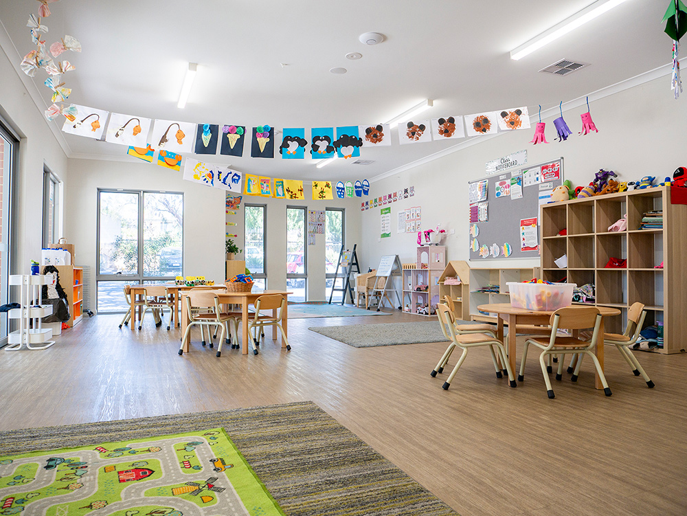 A bright early learning classroom with children's artwork strung across the ceiling, multiple activity tables and chairs, toy shelves, and a play rug