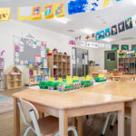 A kindy classroom with children's artwork displayed as bunting across the ceiling, activity tables with egg carton craft projects, toy shelves, and a wooden dollhouse
