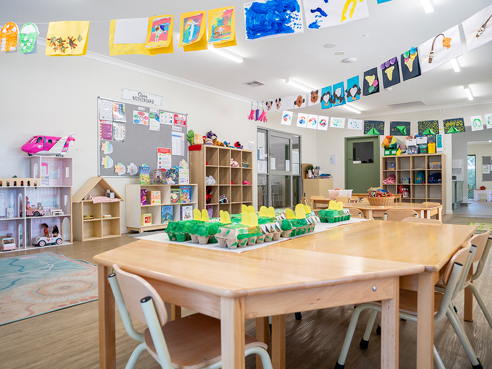 A kindy classroom with children's artwork displayed as bunting across the ceiling, activity tables with egg carton craft projects, toy shelves, and a wooden dollhouse