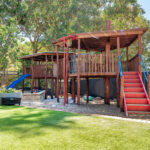 A large two-storey timber palyground with a red staircase, blue slide, and covered deck, set among mature gum trees in a children's outdoor play space