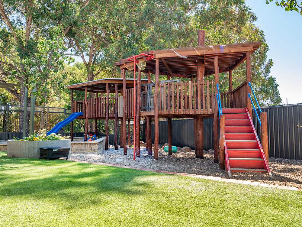 A large two-storey timber palyground with a red staircase, blue slide, and covered deck, set among mature gum trees in a children's outdoor play space