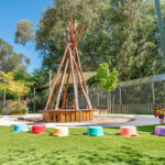 An outdoor play area with a large timber teepee structure surrounded by colourful stepping stools on artificial grass, with shade sails and gum trees in the background
