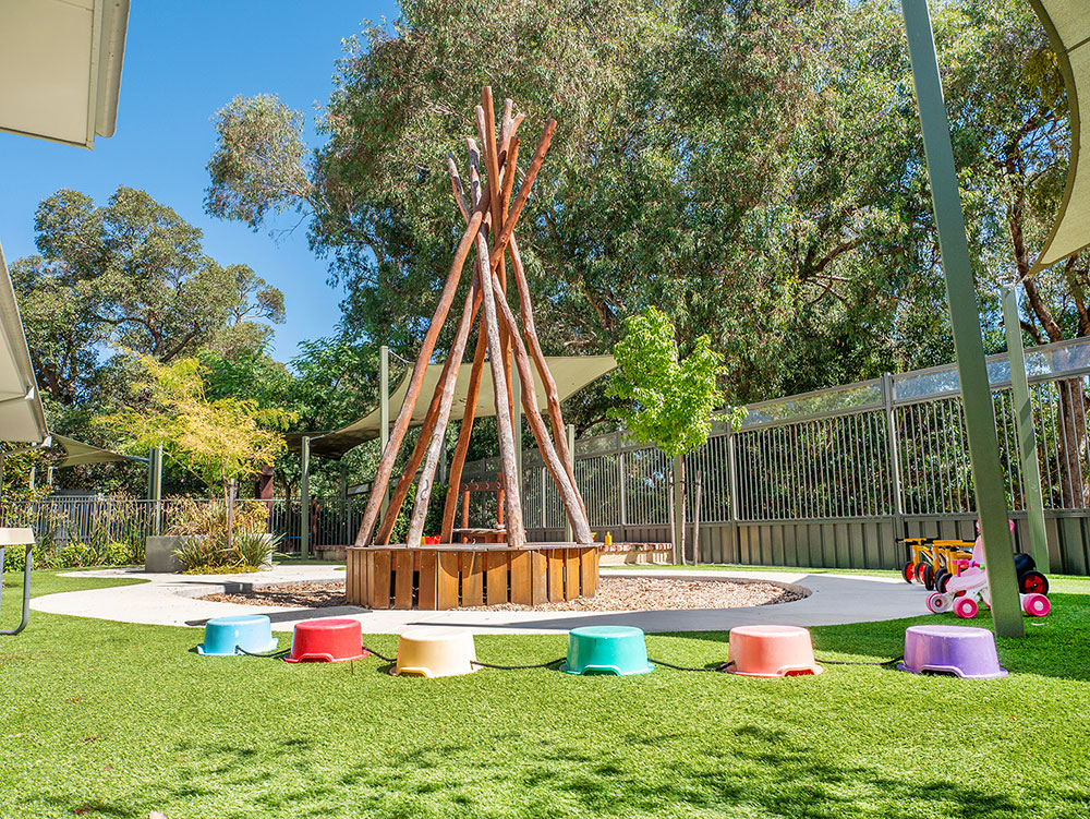 An outdoor play area with a large timber teepee structure surrounded by colourful stepping stools on artificial grass, with shade sails and gum trees in the background