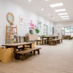 A bright, spacious early learning centre hallway with rustic timber picnic tables and benches, a ladder bookshelf, and an educator noticeboard along the wall