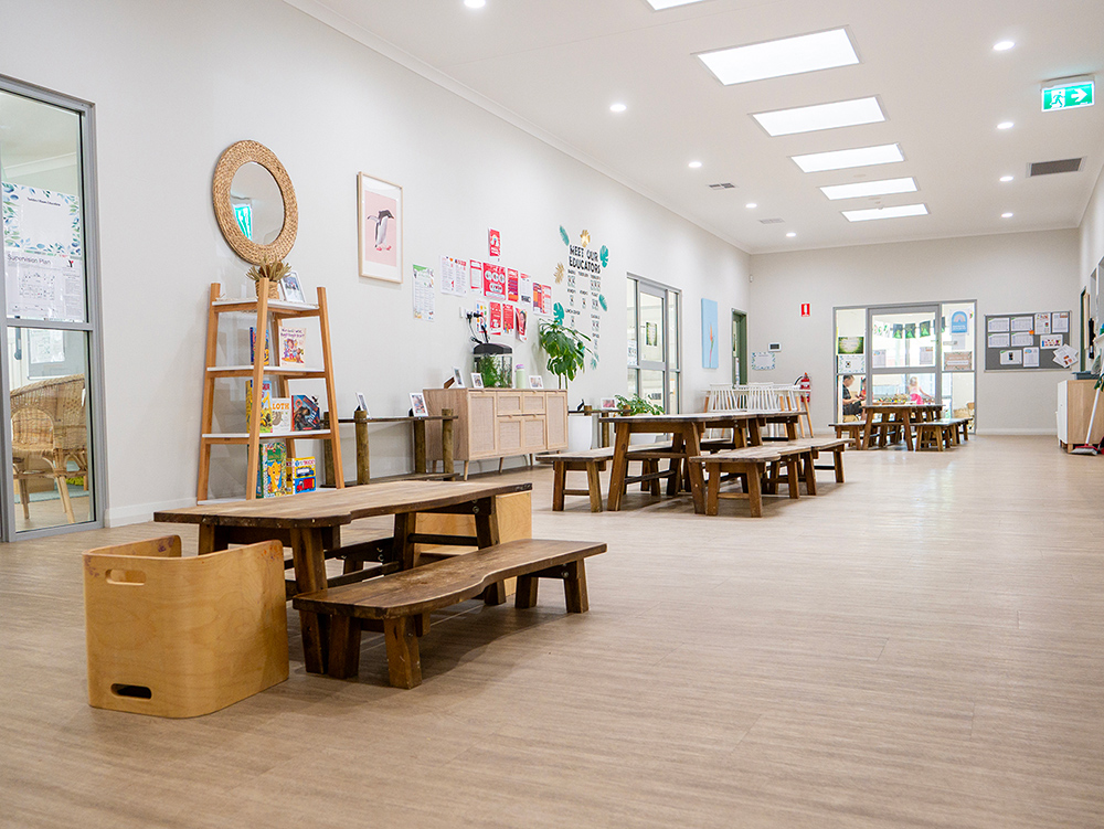 A bright, spacious early learning centre hallway with rustic timber picnic tables and benches, a ladder bookshelf, and an educator noticeboard along the wall