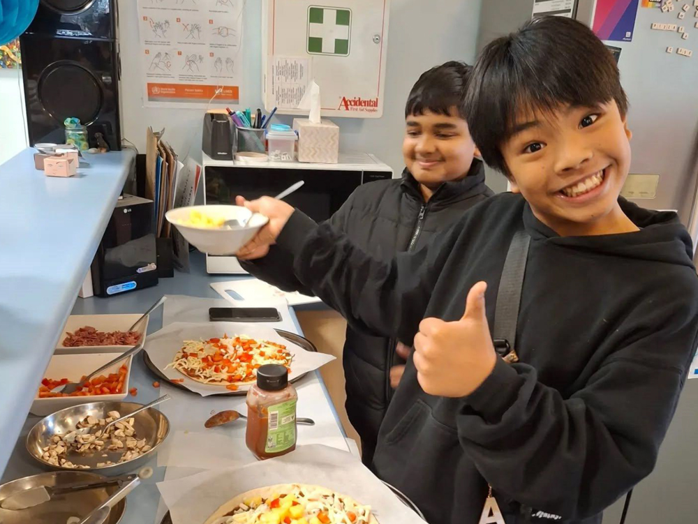 Two young people smiling proudly while making pizza at a bench in the kitchen
