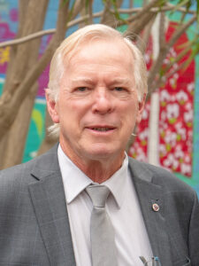 Smiling Y WA Board Member Bruce in a grey suit and tie, photographed outdoors against a colourful mural backdrop
