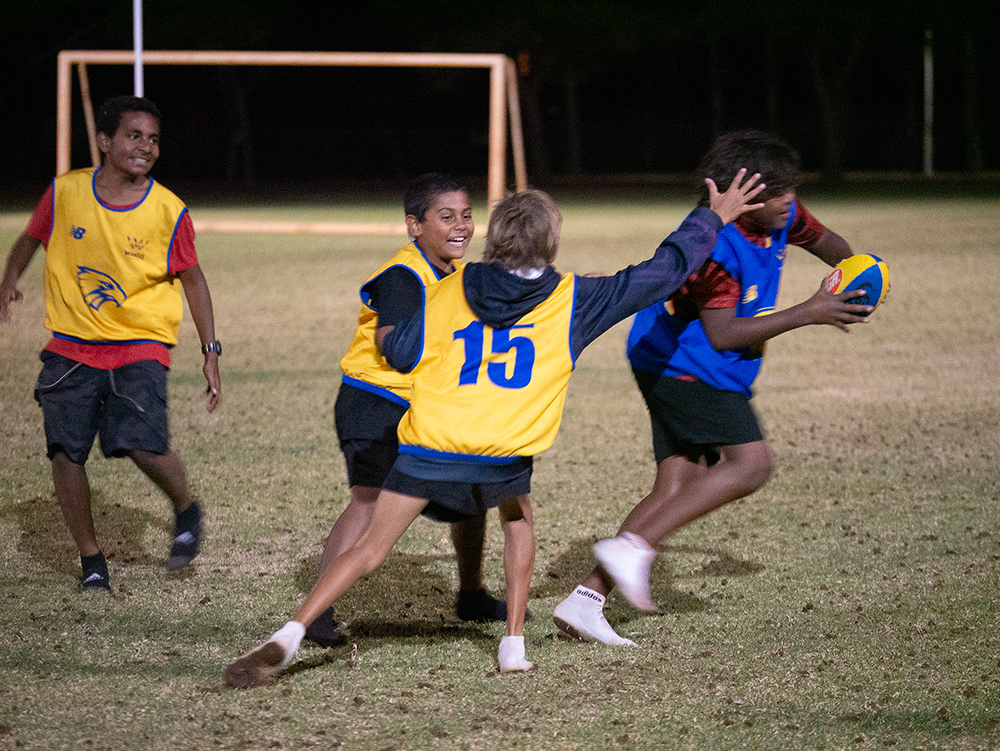 Three children in yellow and blue West Coast Eagles bibs compete for the ball during a night-time AFL game on a grass oval