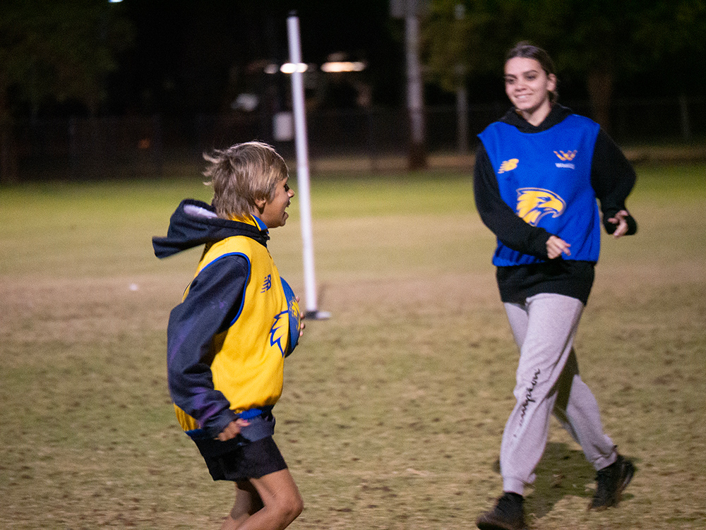 Two young people wearing West Coast Eagles bibs run and laugh together on a lit oval during a night-time AFL activity