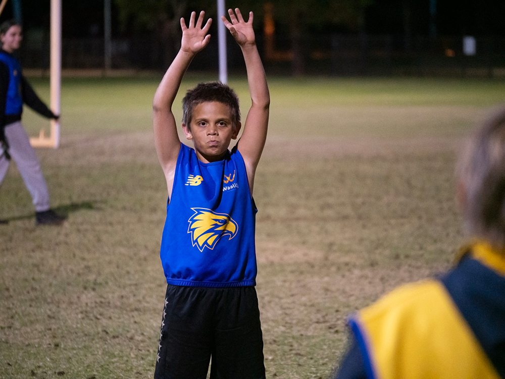 A child in a West Coast Eagles bib raises both hands in the air during an evening AFL skills session on a grass oval