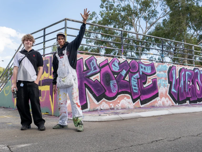 Two young people posing excitedly in front of a wall outside with colourful street art on it