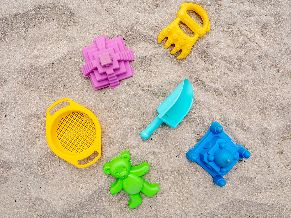 Colourful plastic sand toys including moulds, a sieve, a spade and a rake laid out on sand