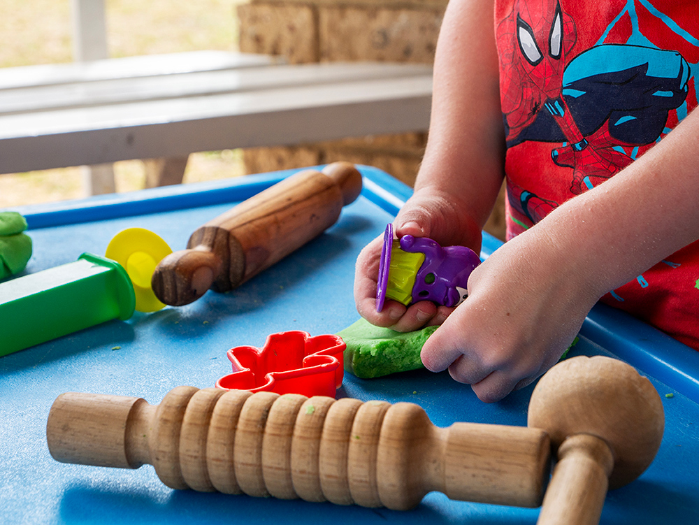 Close-up of a child's hands pressing a purple animal-shaped cutter into green playdough, with wooden rolling pins and shape cutters on a blue tray