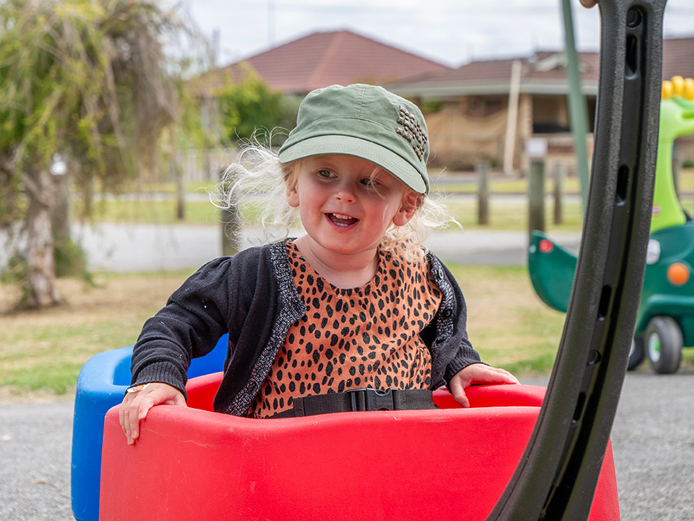 A smiling toddler in a green cap and leopard print top sits in a red and blue ride-on toy at an outdoor play session