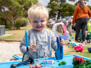 A toddler in a dinosaur jumper plays with playdough and tools at a blue activity table during an outdoor play session