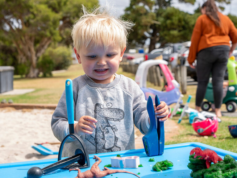 A toddler in a dinosaur jumper plays with playdough and tools at a blue activity table during an outdoor play session