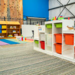 OSHC play area with colourful storage shelving, toys, books, and a rainbow mat, beside an indoor climbing wall.