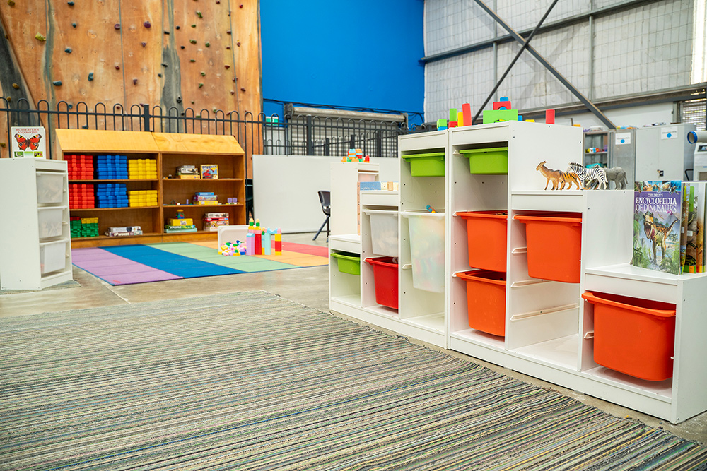OSHC play area with colourful storage shelving, toys, books, and a rainbow mat, beside an indoor climbing wall.