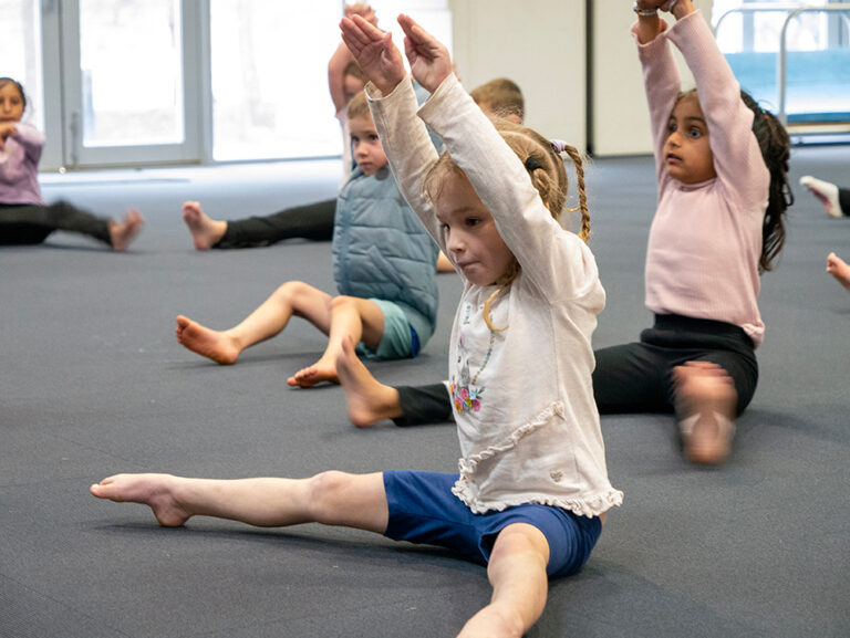 Young children sit on a mat with arms raised during a gymnastics or movement class in a studio