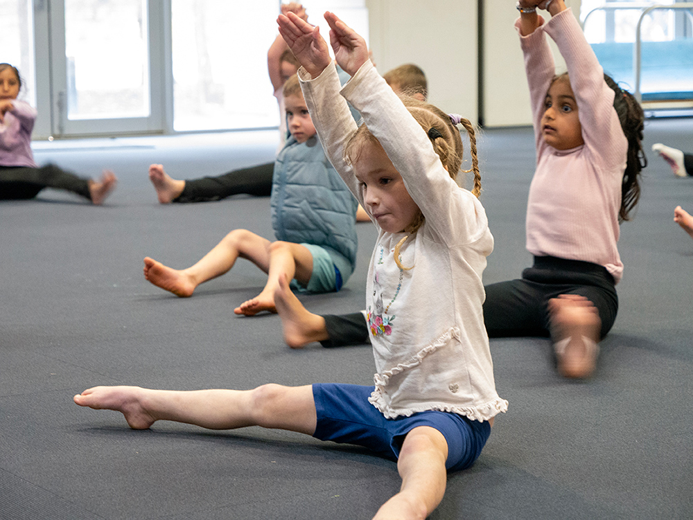 Young children sit on a mat with arms raised during a gymnastics or movement class in a studio