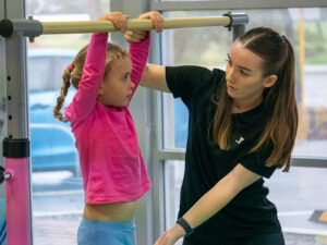 A Y WA gymnastics coach assists a young girl hanging from a bar during a gymnastics class