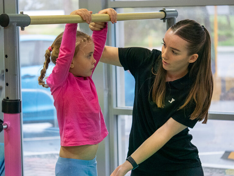 A Y WA gymnastics coach assists a young girl hanging from a bar during a gymnastics class