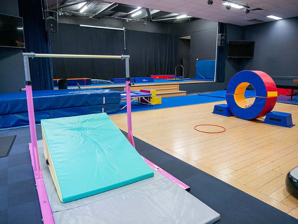 An empty gymnastics studio set up with colourful equipment including a bar, crash mat, foam tunnel, and mini trampoline