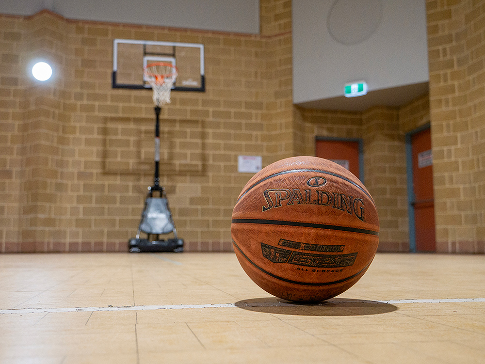 A Spalding basketball sits on a timber court floor in the foreground with a basketball hoop visible in the background