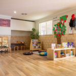 A warm early learning classroom with timber floors, a "Family Friendship" tree display with photos, a children's bookshelf, floor cushions, and Indigenous artwork on the wall