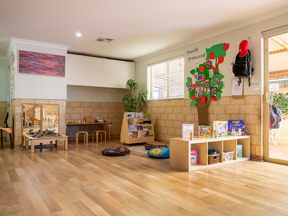 A warm early learning classroom with timber floors, a "Family Friendship" tree display with photos, a children's bookshelf, floor cushions, and Indigenous artwork on the wall