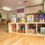 A low timber bookshelf displaying colourful children's books and wicker storage baskets in an early learning centre room