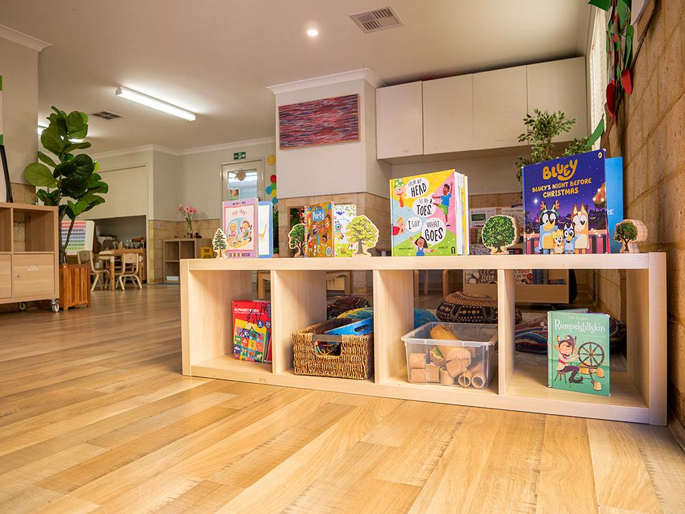 A low timber bookshelf displaying colourful children's books and wicker storage baskets in an early learning centre room