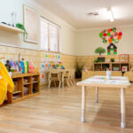 A warm early learning classroom with timber floors, a "Family Friendship" tree display with photos, a children's bookshelf, floor cushions, and Indigenous artwork on the wall