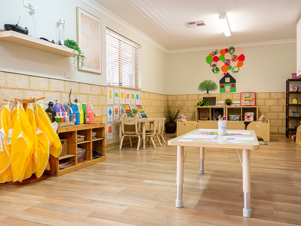 A warm early learning classroom with timber floors, a "Family Friendship" tree display with photos, a children's bookshelf, floor cushions, and Indigenous artwork on the wall
