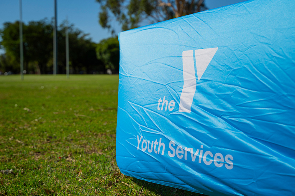 A close-up of a blue Y WA Youth Services branded banner on grass at an outdoor park on a sunny day