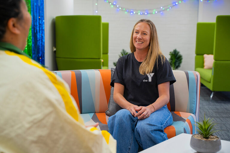 A young person and a Y WA staff member in a black Y t-shirt having a relaxed conversation on a colourful sofa in a vibrant youth centre room